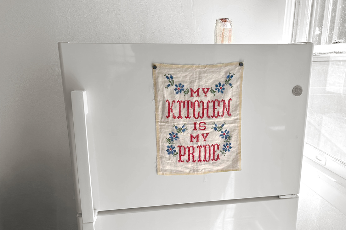 "MY KITCHEN IS MY PRIDE" cross-stitched piece of fabric on front of a freezer.